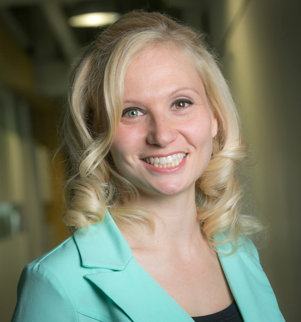 Jana Schaich Borg with blonde curly hair is smiling at the camera. She is wearing a light green blazer over a black top. The background is a softly blurred indoor setting.