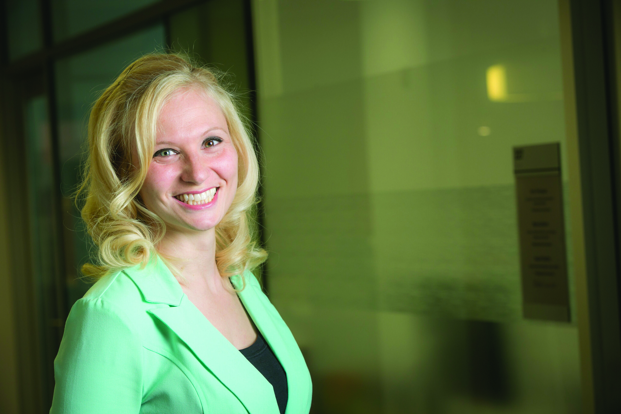 Jana Schaich Borg with long blonde hair smiles while standing indoors. They are wearing a light green blazer over a black top. The background shows a glass wall with an office setting.