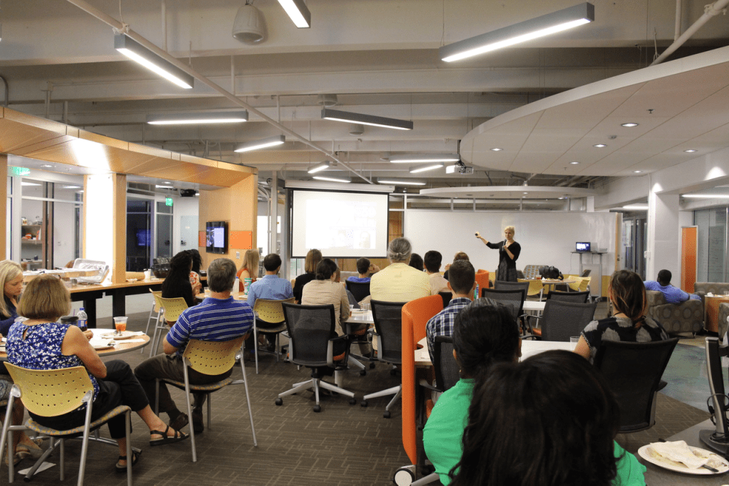 Jana Schaich Borg presents to a group of people seated in a modern conference room. Attendees sit on chairs around tables, facing a large screen. The room has bright lighting, with contemporary decor and an open layout.