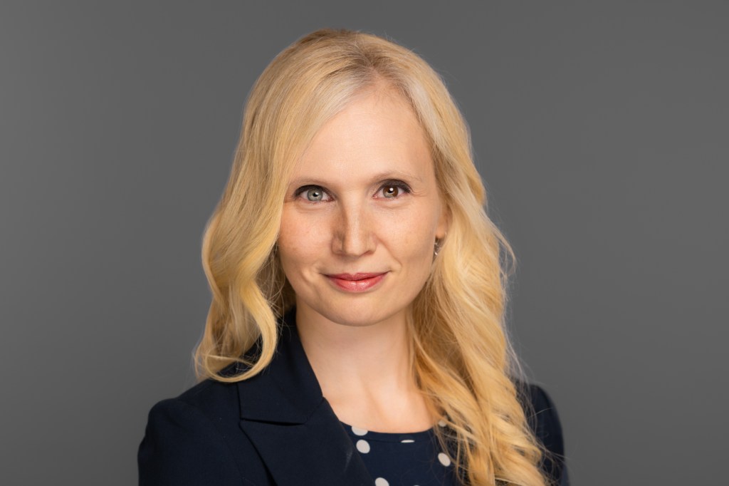 Jana Schaich Borg with long blonde hair wearing a dark blazer and a polka-dot top stands against a plain gray background, looking confidently at the camera.