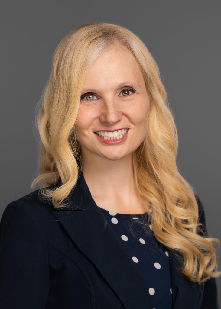 Jana Schaich Borg with long blonde hair smiles in a professional photo,, wearing a navy blue shirt with white polka dots and a dark navy blazer. The background is a plain gray.
