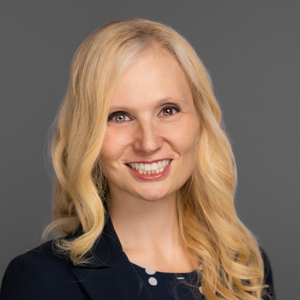 Jana Schaich Borg with long blonde hair smiles in a professional photo, wearing a navy blue shirt with white polka dots and a dark navy blazer. The background is a plain gray.