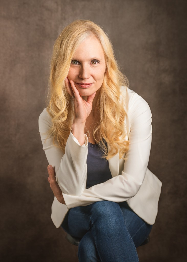 Dr. Jana Schaich Borg with long blonde hair sits on a stool, wearing a white blazer and blue jeans. She rests her chin on her hand and smiles softly at the camera against a brown textured background.