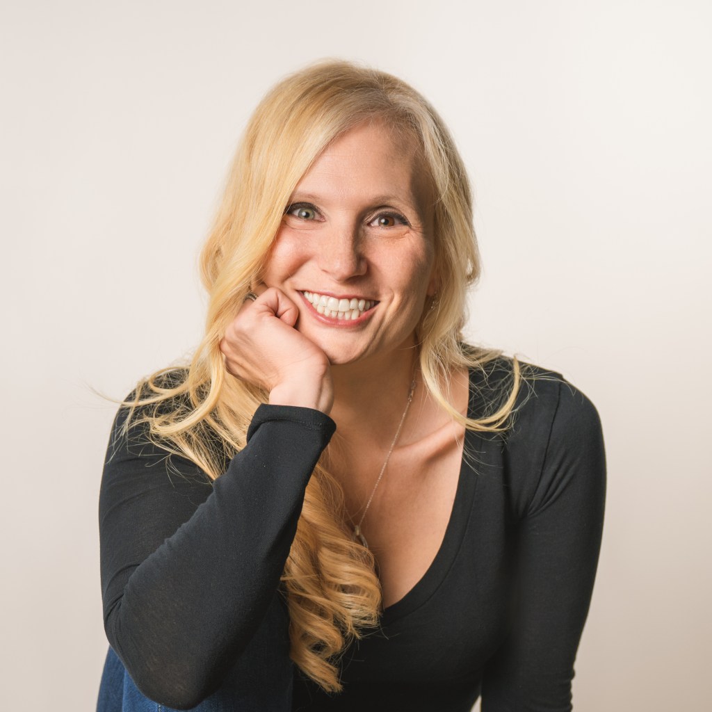 Jana Schaich Borg with long blonde hair, wearing a black top, smiles broadly at the camera. She rests her chin on her hand, sitting against a neutral background.