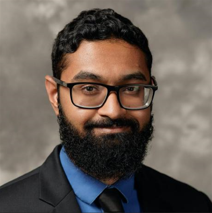 Gayan with a beard and glasses, wearing a black suit with a blue shirt and black tie, smiles slightly. The background is a soft, abstract gray.