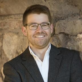 Gus with glasses and a beard stands against a stone wall, wearing a dark blazer and a white shirt. His arms are crossed, and he is smiling at the camera.